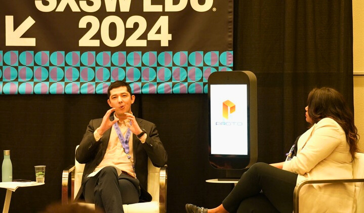A panel discussion at SXSW EDU 2024, with two participants on stage. On the left, a man in a black blazer is making a hand gesture, possibly signing, indicative of a discussion about accessibility or technology. To the right, a woman is seated looking towards him, attentively. They are both in front of a backdrop that reads 'SXSW EDU 2024' with a pattern of cyan circles on a black field. Beside them is a digital display stand showcasing the 'Proto' logo, hinting at a technology or educational presentation.